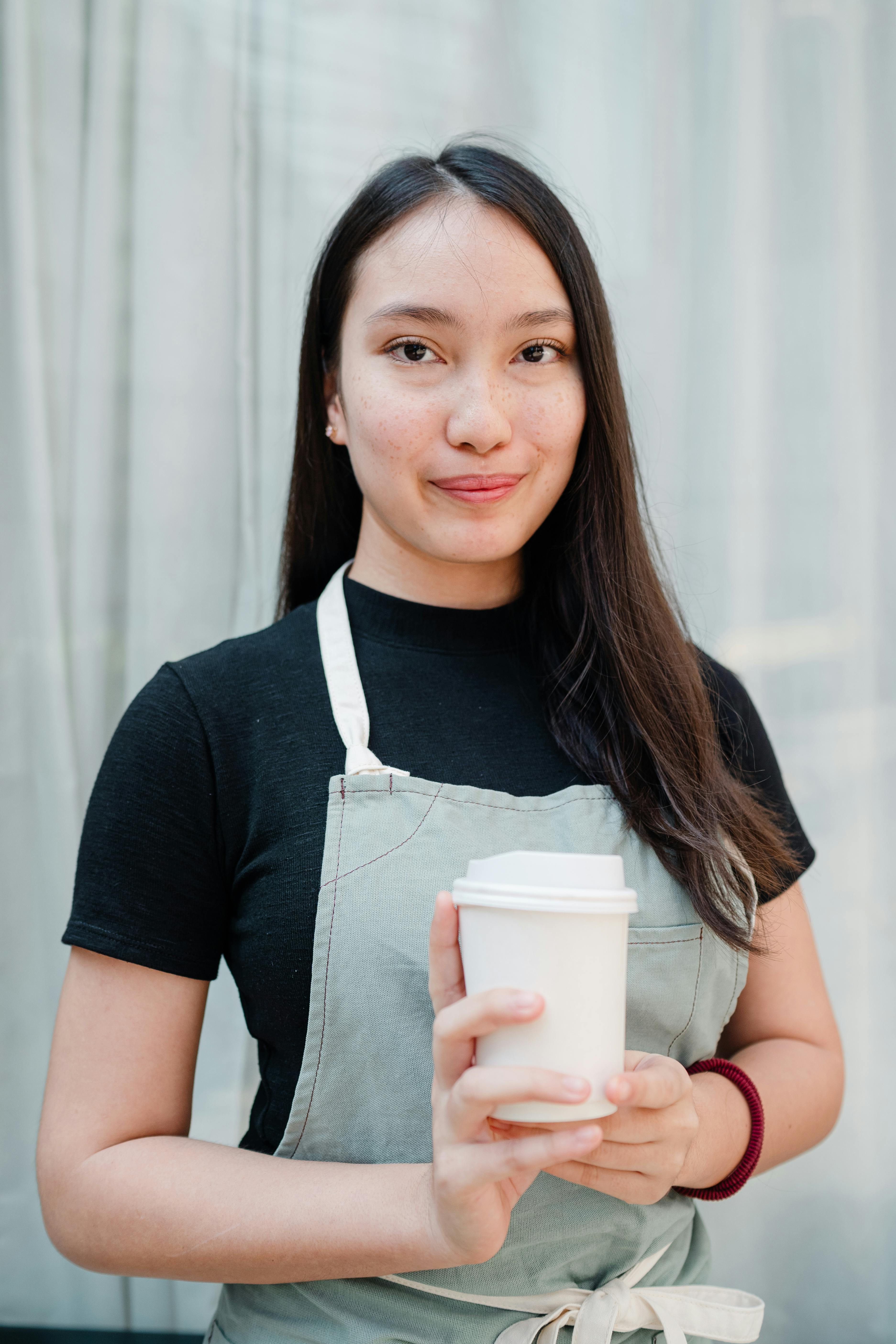 smiling Asian woman with white takeaway paper cup in hands standing against light blue curtains in cozy cafe and looking at camera
